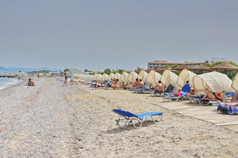 Kos - Psalidi Strandabschnitt mit Kies und Sand nahe Kos-Stadt. Zahlreiche Liegen mit halbrunden Sonnenschirmen stehen in einer Reihe, dahinter sieht man Hotelgebäude mit dem Schriftzug 'Ramira Beach'. Menschen sonnen sich und genießen das Meer.