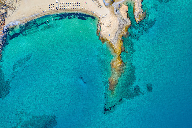 Luftaufnahme von Potamos Beach mit Sandstrand, Sonnenschirmen und felsiger Landzunge im türkisfarbenen Meer