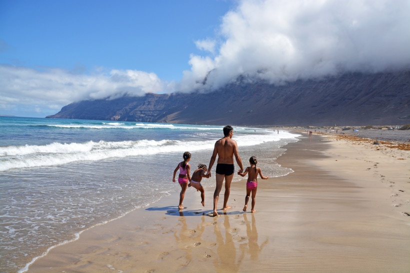 Familienurlaub auf Lanzarote – Strandtag mit Kindern an der Playa de Famara Vater mit drei Kindern beim Spielen und Spazieren am Strand von Lanzarote, mit Blick auf die Klippen der Playa de Famara – ideal für Familienurlaub am Meer.