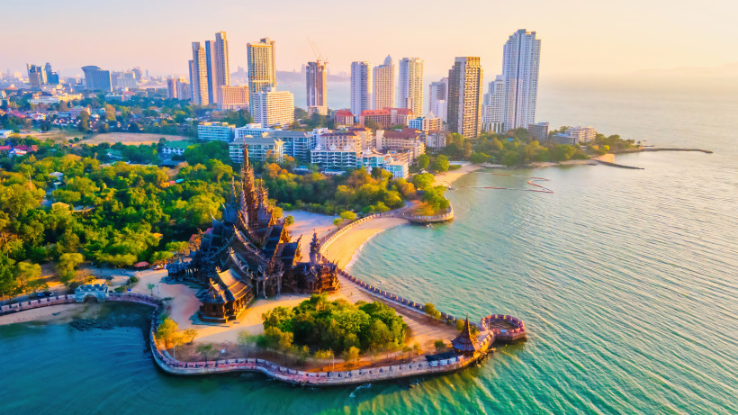 Thailand Luftaufnahme einer kunstvoll geschnitzten Tempelanlage am Meer, umgeben von grüner Vegetation. Im Hintergrund ragen moderne Hochhäuser einer Küstenstadt in den Himmel. Die Abendsonne taucht die Szene in warmes Licht.