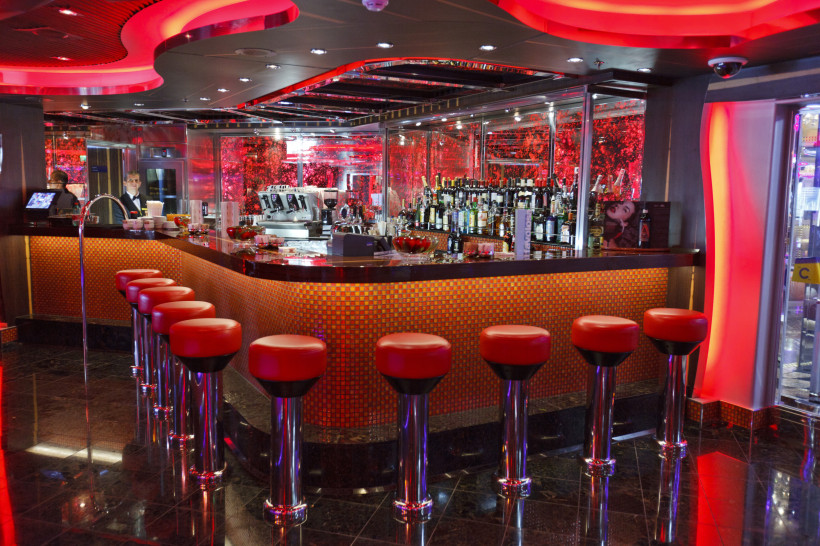 Costa Favolosa – Camelot Piano Bar, Barbereich mit roten Barhockern und beleuchteter Theke Barbereich der Camelot Piano Bar auf der Costa Favolosa mit roter Beleuchtung, Barhockern und Flaschenregalen