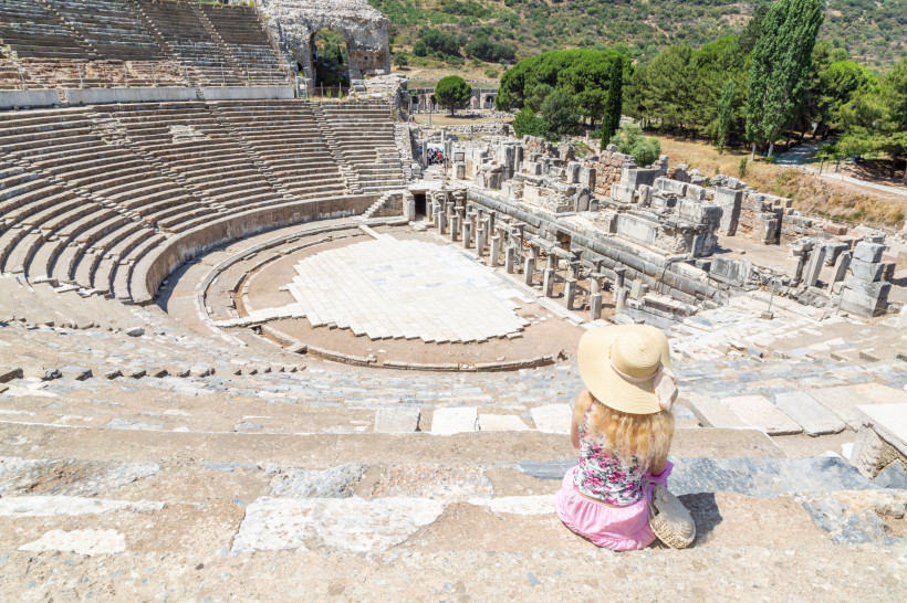 Eine Frau mit Sonnenhut und Sommerkleidung sitzt in den oberen Rängen eines antiken Amphitheaters in Ephesos, Türkei. Sie blickt auf die halbkreisförmige Bühne mit umliegenden Ruinen und grüner Landschaft im Hintergrund.