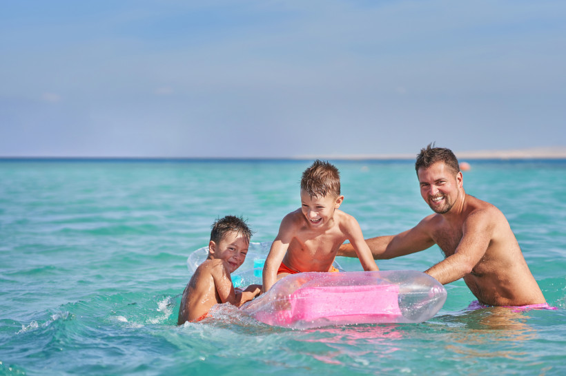 Ägypten - Hurghada Ein Vater spielt mit zwei kleinen Jungen lachend im türkisfarbenen Meer. Die Kinder sitzen auf einer aufblasbaren pinken Luftmatratze. Im Hintergrund ist der weite Ozean zu sehen, die Stimmung ist fröhlich und unbeschwert.