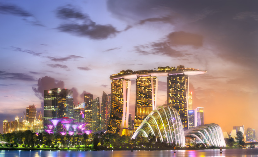 Singapur Blick auf die nächtliche Skyline von Singapur mit dem ikonischen Marina Bay Sands Hotel, das mit seiner markanten Dachplattform über drei Türmen beleuchtet ist. Im Vordergrund leuchten die futuristischen Gewächshäuser der Gardens by the Bay in sanften Far