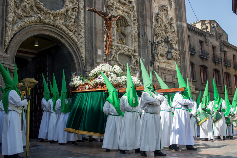 Religiöse Prozession vor einer kunstvoll verzierten Kirche, bei der Männer in weißen Gewändern und grünen Spitzhauben eine Statue von Jesus am Kreuz auf einem blumengeschmückten Tragegestell tragen