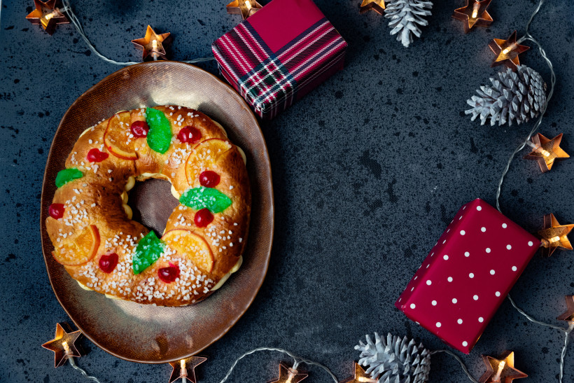 Ein traditioneller spanischer Weihnachtskuchen in Kranzform liegt auf einem braunen Teller. Der Kuchen ist mit kandierten Früchten, Zucker und Orangenscheiben dekoriert. Daneben stehen zwei bunt verpackte Geschenke. Die Szenerie ist von Tannenzapfen, Ster