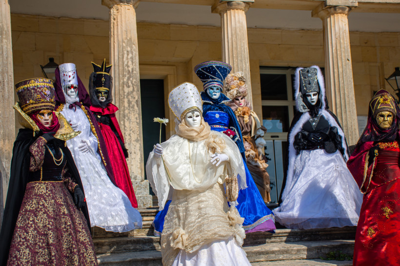 Karneval auf Korfu: Eine Gruppe kunstvoll maskierter Menschen steht in prächtigen Gewändern vor einem neoklassizistischen Gebäude. Die aufwendigen Kostüme erinnern an den venezianischen Karneval und unterstreichen die kulturelle Verbindung der Insel mit I