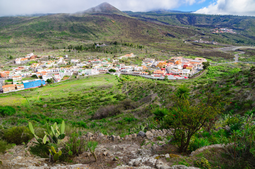 Santiago del Teide – Bergpanorama und grünes Tal Weite Berglandschaft mit grünem Tal und dem Dorf Santiago del Teide auf Teneriffa, Häuser mit roten Dächern inmitten von Feldern und Hügeln, Berge im Hintergrund und Wolken über den Gipfeln.