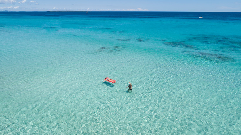 Luftaufnahme von türkisfarbenem, klarem Meer vor Formentera mit zwei Personen im flachen Wasser, eine auf einer Luftmatratze