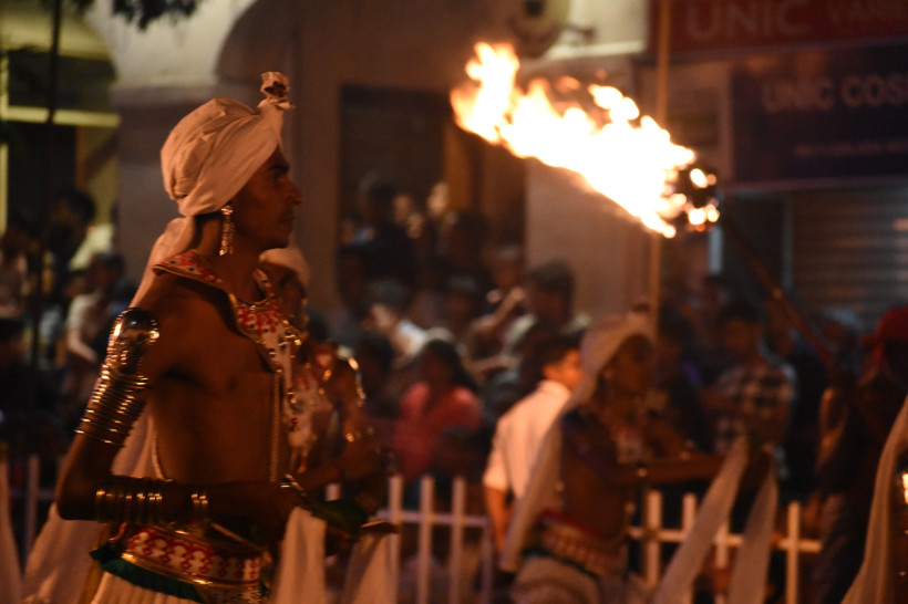 Ein traditionell gekleideter Mann trägt bei Nacht eine große, brennende Fackel während einer Prozession. Er trägt weiße Kleidung mit Schmuckelementen und eine Kopfbedeckung. Im Hintergrund sind Zuschauer und weitere Teilnehmer der Parade zu sehen.