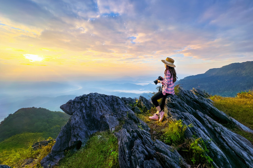 Eine Frau mit Kamera sitzt auf einem Felsen und blickt beim Sonnenaufgang über eine weite Berglandschaft mit Nebel in den Tälern. Sie trägt Wanderschuhe, Strohhut und kariertes Hemd.