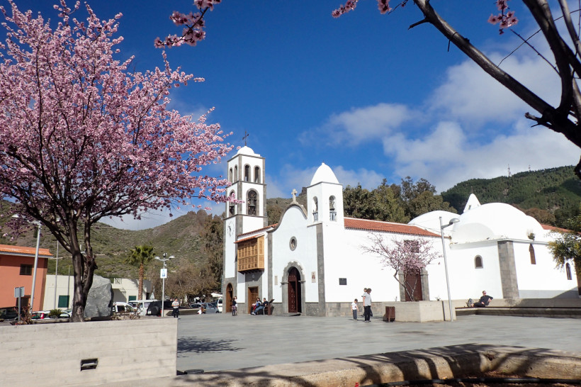 Santiago del Teide – Traditionelle Kirche und Mandelblüte im Bergdorf Weiße Dorfkirche mit zwei Glockentürmen in Santiago del Teide auf Teneriffa, vorne blühende rosa Mandelbäume und dahinter grüne Berghänge unter blauem Himmel.