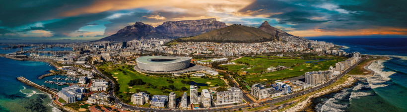 Südafrika Panoramaaufnahme von Kapstadt in Südafrika mit dem markanten Tafelberg im Hintergrund. Im Vordergrund ist das Cape Town Stadium gut erkennbar, daneben weitläufige Grünflächen, die Innenstadt und die Waterfront mit dem Hafen. Rechts im Bild verläuft die Kü