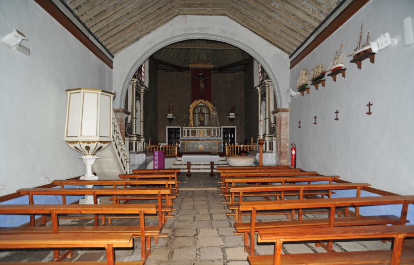 Lanzarote Innenraum einer kleinen Kirche mit Holzbänken, Kanzel, Altar und Kreuzigungsdarstellung. An der rechten Wand hängen Modelle von Segelschiffen auf kleinen Regalen.