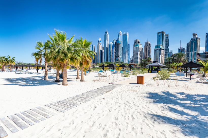 Dubai Heller Sandstrand in Dubai mit Palmen, Sonnenliegen und schattigen Pavillons. Im Hintergrund erhebt sich die moderne Skyline von Dubai Marina mit zahlreichen futuristischen Hochhäusern und Wolkenkratzern. Der Himmel ist strahlend blau, die Szene wirkt son