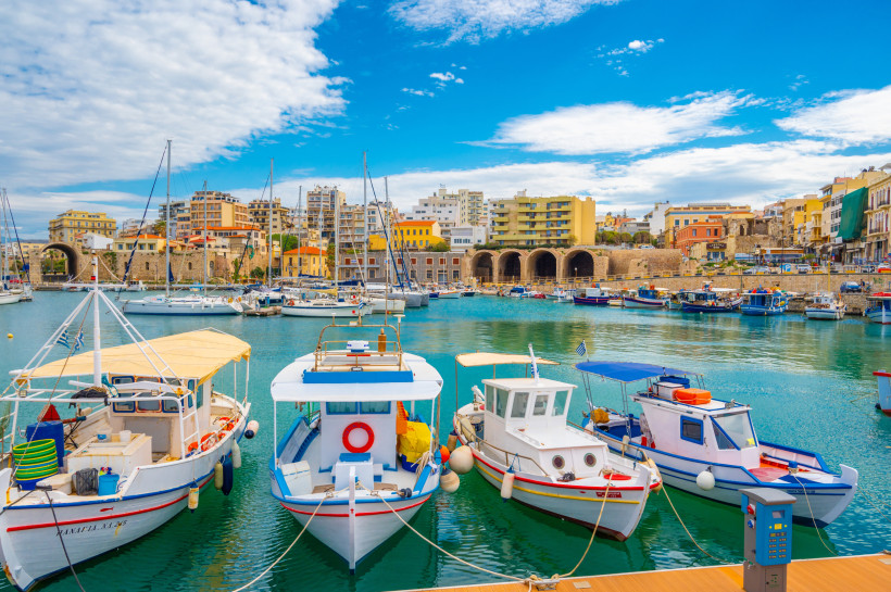 Venezianischer Hafen in Heraklion mit kleinen Booten im Vordergrund, Hafenmauer und Stadtbebauung unter blauem Himmel