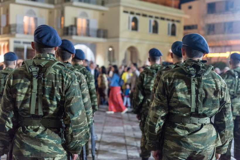 Kreta Mehrere griechische Soldaten in Tarnuniformen und blauen Baretts stehen mit dem Rücken zur Kamera bei einer offiziellen Veranstaltung oder Zeremonie. Im Hintergrund versammeln sich Zuschauer vor einem Gebäude mit Balkonen und Bögen, vermutlich auf einem ö
