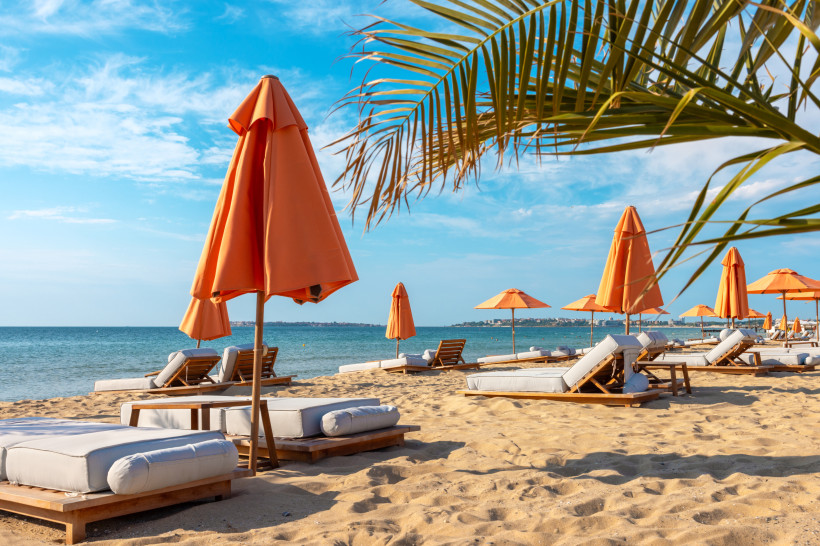 Bulgarien Sonnige Strandlandschaft mit bequemen weißen Liegen und orangefarbenen Sonnenschirmen auf goldenem Sand. Im Hintergrund das ruhige, blaue Meer und eine kleine Stadt an der Küste. Eine Palme ragt ins Bild und verstärkt die Urlaubsatmosphäre