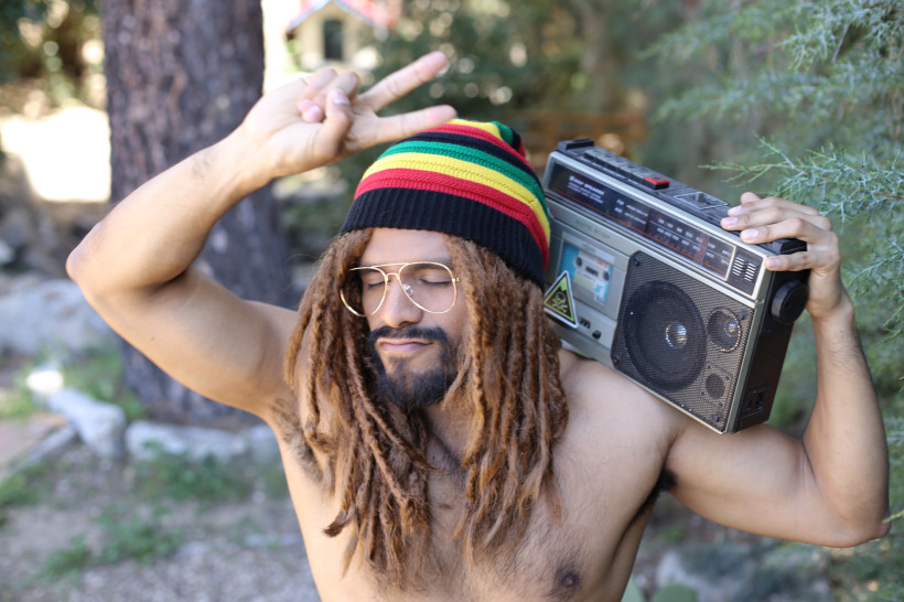 Jamaika Ein junger Mann steht oberkörperfrei draußen im Grünen. Er trägt eine Mütze in den Rastafari-Farben Rot, Gelb, Grün und Schwarz sowie eine auffällige Brille. Seine Haare sind zu Dreadlocks gestylt. Auf der Schulter trägt er ein großes, altmodisches Radio.