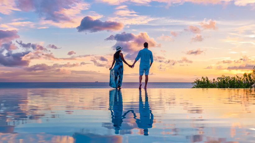 Romantik pur beim Sonnenuntergang auf Gran Canaria in Paar hält sich an den Händen und genießt den Sonnenuntergang am Infinity-Pool mit Blick auf das Meer. Die sanften Farben des Himmels spiegeln sich im Wasser und schaffen eine traumhafte Atmosphäre.