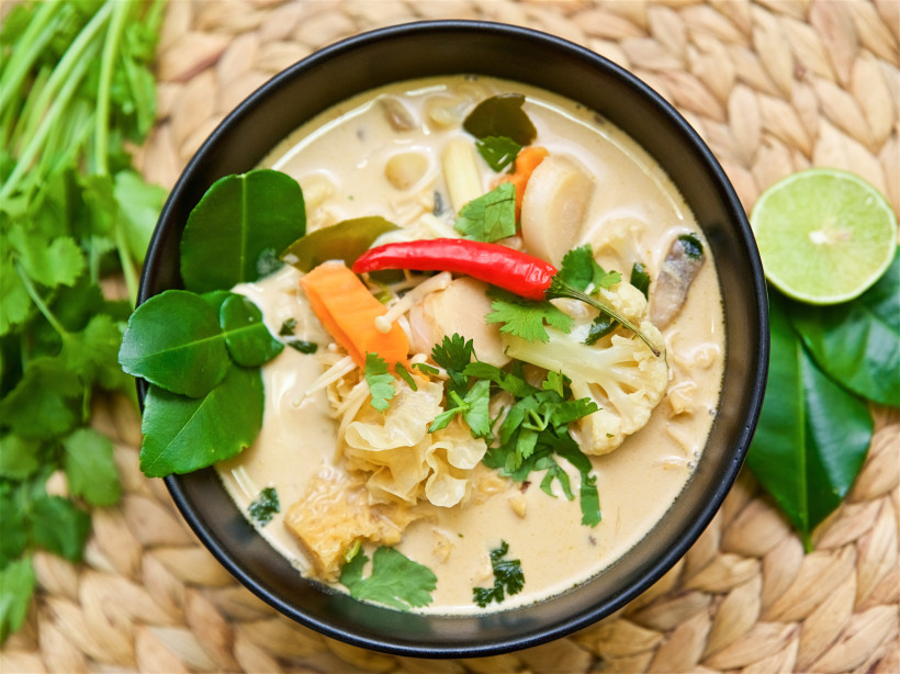 Thailand - Krabi Das Bild zeigt eine schwarze Schale mit einer cremigen thailändischen Kokossuppe (vermutlich Tom Kha). Die Suppe ist mit frischen Zutaten wie Blumenkohl, Karotten, Pilzen, Koriander und einer ganzen roten Chilischote dekoriert. Um die Schüssel herum liege