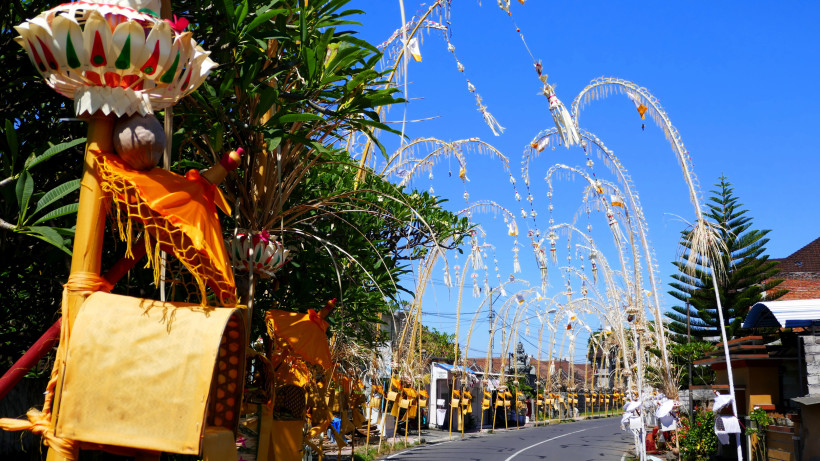 Bali Eine Straße auf Bali ist mit kunstvoll verzierten Penjor-Stäben geschmückt. Die hohen Bambusstangen mit hängenden Opfergaben und bunten Stoffen biegen sich in Richtung Straße. Links im Vordergrund sind reich dekorierte Altäre mit orangefarbenen Tüchern zu