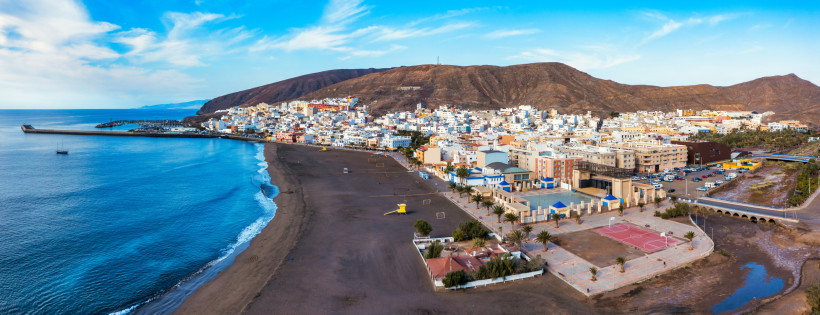 Panoramaaufnahme von Gran Tarajal auf Fuerteventura mit weitem Stadtstrand, Hafen und der Kulisse brauner Berge im Hintergrund.