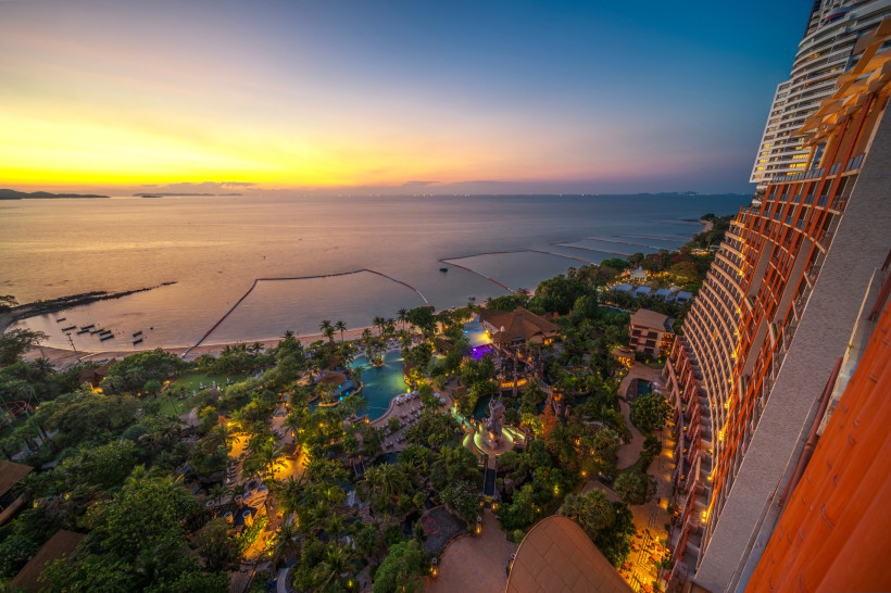 Thailand Pattaya Blick vom Hochhaus auf ein beleuchtetes Luxusresort mit tropischer Gartenanlage und Pools direkt am Strand von Pattaya. Im Hintergrund ruhiges Meer bei Sonnenuntergang, der Himmel leuchtet in Gelb- und Blautönen.
