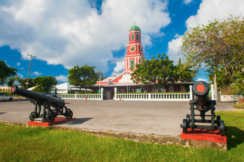 Barbados Historisches Gebäude mit rotem Uhrenturm und grüner Kuppel in Bridgetown, Barbados. Vor dem Gebäude stehen zwei alte schwarze Kanonen auf rot markierten Sockeln. Der Platz ist von einer weißen Balustrade eingefasst, umgeben von Bäumen und unter einem Himm