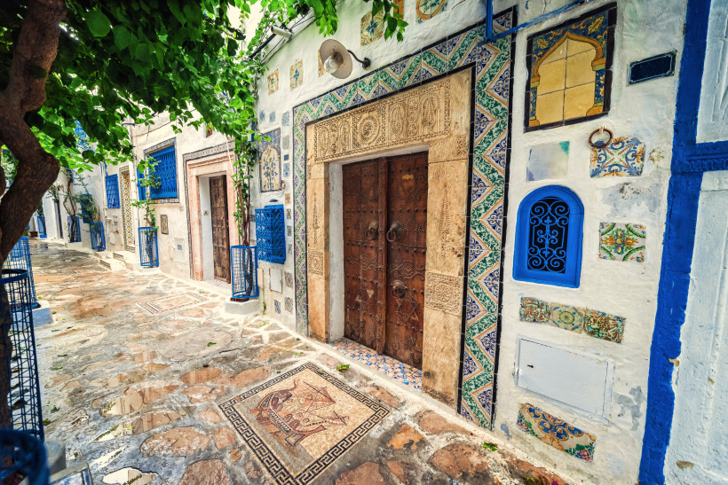 Eine mit Mosaiken verzierte Gasse in der Medina von Tunis. Links ein alter Baum, rechts eine Hausfassade mit kunstvollen Verzierungen, bunten Fliesen und einer massiven Holztür. Die Straße ist gepflastert und mit dekorativen Mosaiken versehen.