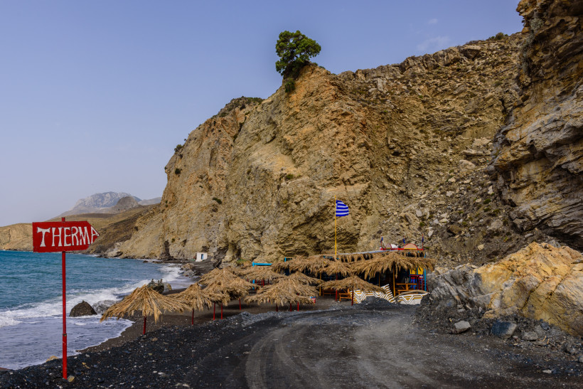 Kos - Therma Eingang zum Strand Therma auf Kos mit Felsen, Strohdächern und einer griechischen Flagge im Wind