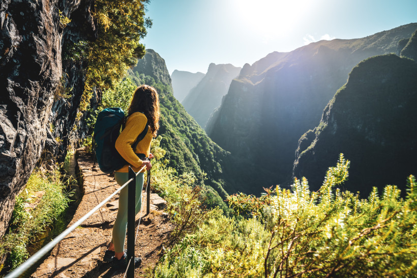 Madeira Eine Frau mit Rucksack steht auf einem gesicherten Pfad an einer bewässerten Levada. Vor ihr öffnen sich     dicht bewaldete Hänge und zerklüftete Berge, die Sonne strahlt durch leichten Dunst ins Tal.