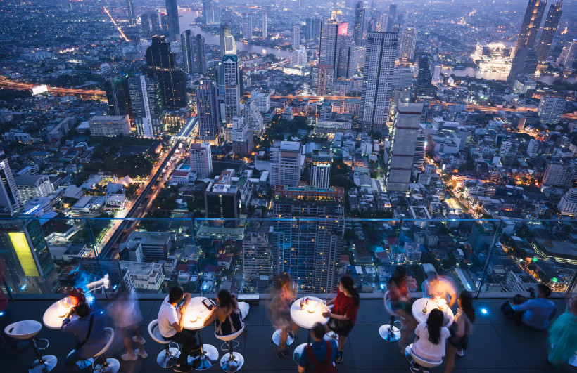 Bangkok bei Nacht vom Rooftop – Spektakuläre Aussicht über die Stadt aus luftiger Höhe Menschen genießen den Blick auf das nächtliche Bangkok von einer Rooftop-Bar mit Glasgeländer und Tischen