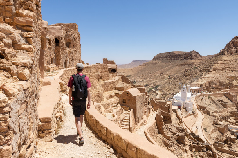 Ein Tourist mit Rucksack und Kappe geht auf einem schmalen Weg durch die Ruinen einer historischen Wüstenstadt aus Lehmstein. Rechts unten ist ein weißes Gebäude mit Türmen zu sehen, das sich von der sonst ockerfarbenen Landschaft abhebt. Im Hintergrund e