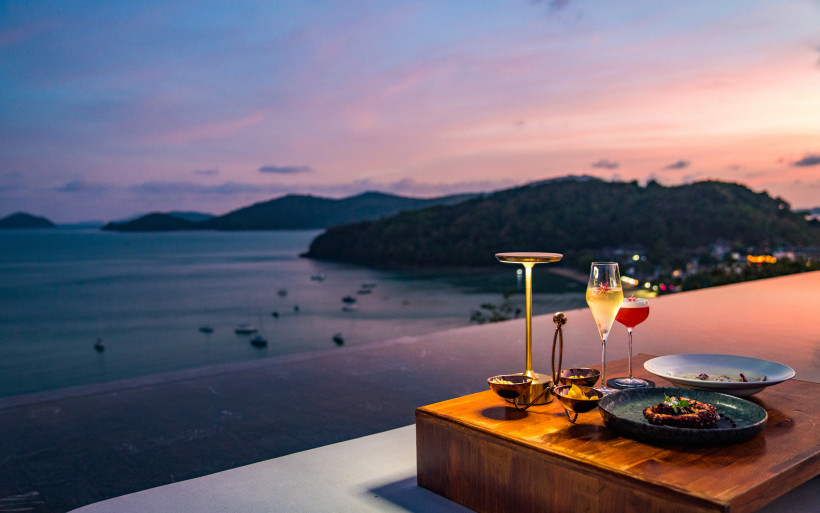 Thailand - Phuket Abendstimmung über dem Meer auf Phuket. Im Vordergrund ein elegant gedeckter Tisch mit modernen goldenen Lampen, einem Glas Champagner, einem Cocktail sowie kunstvoll angerichteten Speisen. Im Hintergrund die Bucht und sanft beleuchtete Hügel unter einem