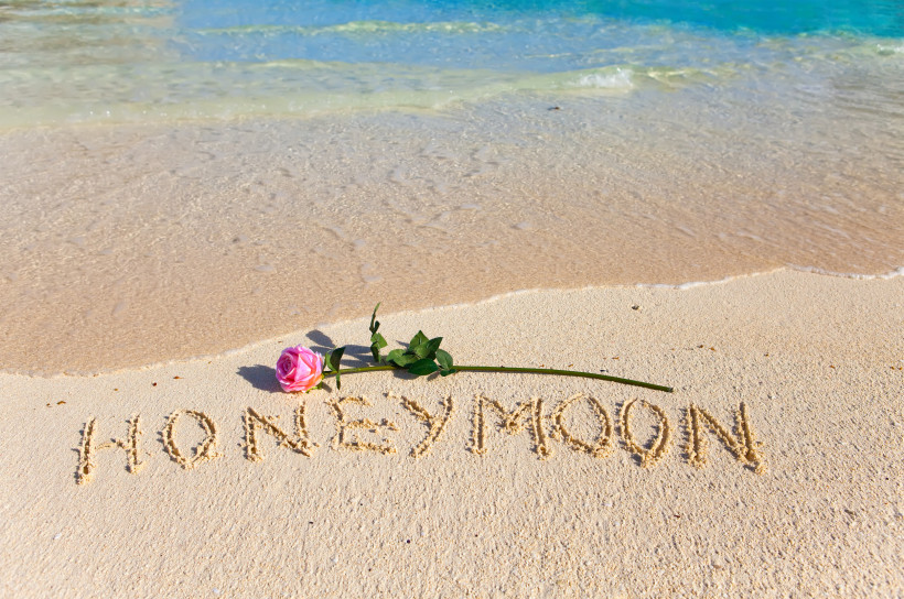 Honeymoon am Meer – romantische Strandkulisse Rose liegt am Strand neben dem Wort „Honeymoon“, das im Sand geschrieben ist. Symbolische Szene für romantische Flitterwochen am Meer.