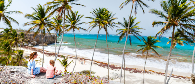 Barbados Zwei Frauen – eine Erwachsene und ein Kind – sitzen auf einem Felsen mit Blick auf einen palmengesäumten Strand und das türkisblaue Meer. Die Szene ist von oben aufgenommen und zeigt Palmen, Felsen, Wellen und eine naturbelassene Küstenlandschaft bei hell