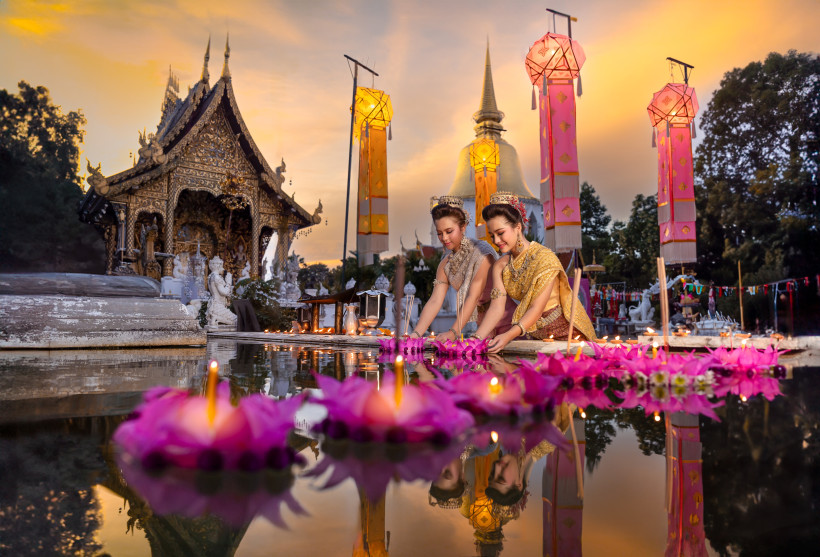 Thailand - Krabi Das Bild zeigt zwei thailändische Frauen in prachtvoller traditioneller Kleidung, die an einem festlich beleuchteten Tempel in der Dämmerung schwimmende Lichter und Blütenkränze ins Wasser setzen – sogenannte Krathongs. Im Hintergrund steht ein kunstvoll