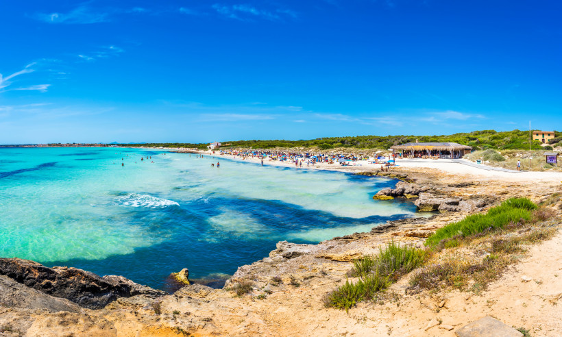 Mallorca - Ses Covetes Eine weite Strandbucht auf Mallorca mit feinem, hellem Sand und türkisfarbenem Wasser. Der Strand ist von einer großen Menschenmenge belebt, die das Meer und die Sonne genießen. Im Hintergrund erstreckt sich ein dichter Pinienwald, der für natürlichen Sch