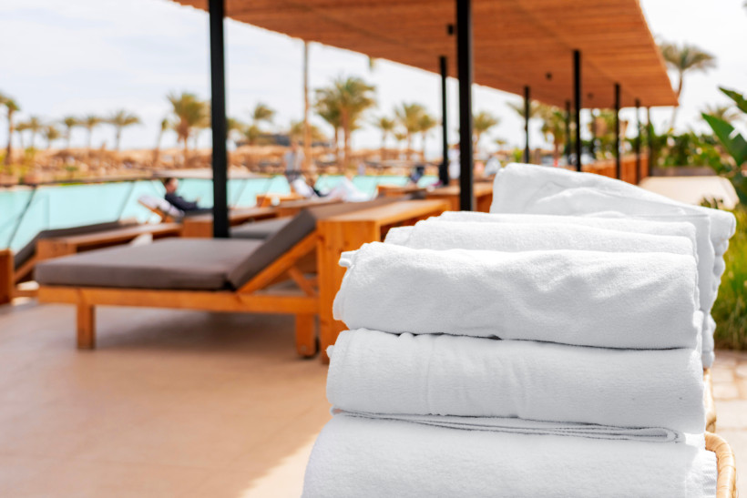 Ägypten - El Gouna Gestapelte weiße Poolhandtücher im Vordergrund, dahinter stilvolle Holzliegen mit Auflagen unter einem schattenspendenden Dach am Pool eines Resorts. Im Hintergrund Palmen und entspannte Gäste.