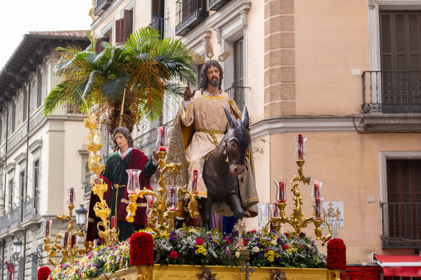 Eine festliche Prozession zeigt eine Figur von Jesus auf einem Esel, begleitet von einer weiteren Figur, auf einer prächtigen Plattform mit Palmen, Blumen und goldenen Kerzenhaltern. Die Jesusfigur trägt ein helles Gewand mit goldener Stickerei und hat go