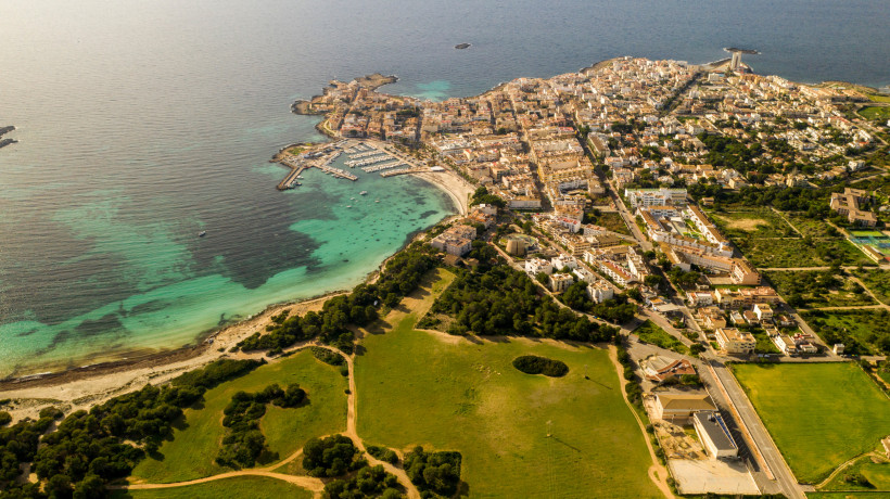 Mallorca - Colònia de Sant Jordi Luftaufnahme einer Küstenstadt auf Mallorca mit einem langen Sandstrand und türkisfarbenem Wasser. Im Vordergrund ist eine weitläufige grüne Landschaft mit Feldern und Bäumen zu sehen. Die Stadt liegt direkt am Wasser und hat einen kleinen Yachthafen. Das