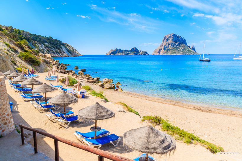 Ibiza Sonniger Sandstrand mit strohgedeckten Sonnenschirmen und blauen Liegestühlen auf der Baleareninsel Ibiza. Das ruhige, türkisfarbene Meer liegt direkt vor der Strandkulisse, im Hintergrund ragen felsige Klippen empor. Im Wasser liegen einige Segelboote vo