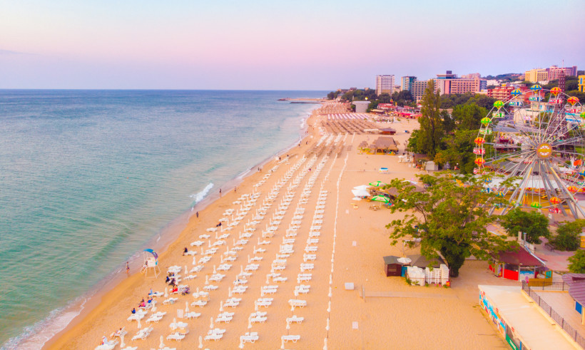 Luftaufnahme des Goldstrands bei Sonnenuntergang. Reihenweise weiße Sonnenliegen und Schirme säumen den langen Sandstrand. Rechts steht ein buntes Riesenrad vor Hotels und Freizeitangeboten. Das Meer schimmert in zarten Blau- und Rosatönen.