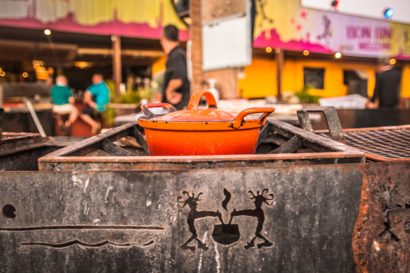 Ein oranger Gusseisentopf steht auf einem traditionellen Grill im Freien, eingerahmt von Metall mit geschnitzten Symbolen. Im Hintergrund Menschen und eine farbenfrohe Kulisse – eine karibische Straßenszene mit lebendiger Atmosphäre.
