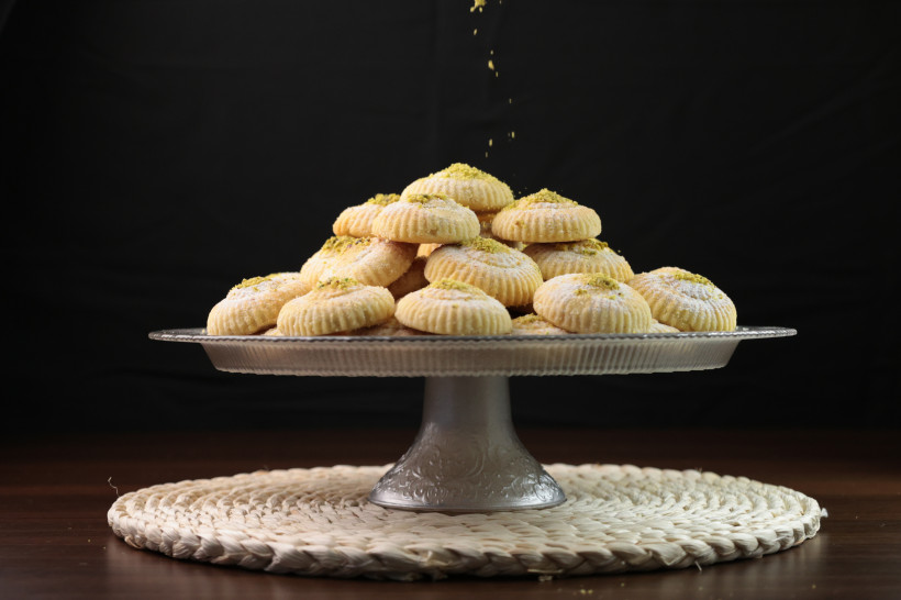 Ägypten - Sharm el Sheikh Ein silberfarbenes Tortenpodest voller traditioneller Maamoul-Kekse, die mit Puderzucker und gehackten Pistazien bestreut sind. Die Kekse sind rund, leicht geriffelt und gestapelt. Der Hintergrund ist schwarz, der Tisch aus Holz, unter dem Podest liegt ei