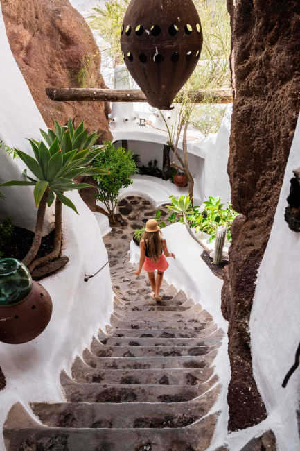 Lanzarote Frau mit Strohhut und Sommerkleid geht eine steinerne Treppe hinunter, umgeben von weiß verputzten Wänden, Pflanzen und Felsen in organisch geschwungener Architektur