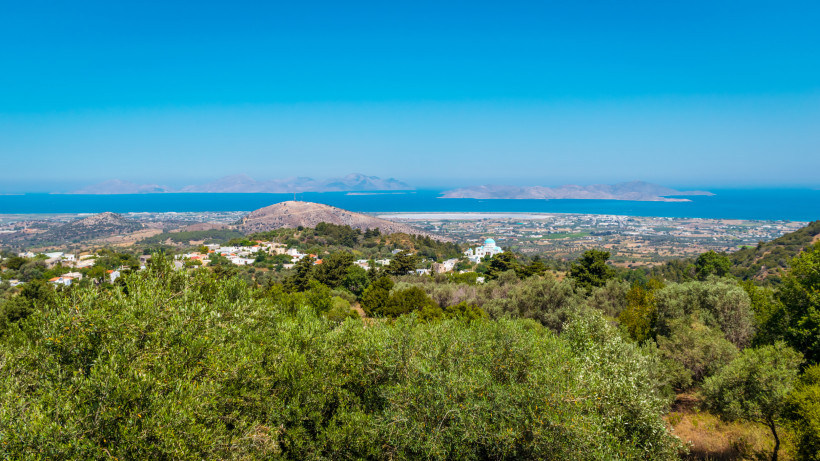 Kos Panoramablick von einem Hügel über die grüne Insel Kos mit ihren verstreuten Häusern, Olivenbäumen und Feldern. Im Hintergrund das tiefblaue Meer mit weiteren Inseln am Horizont unter wolkenlosem Himmel.