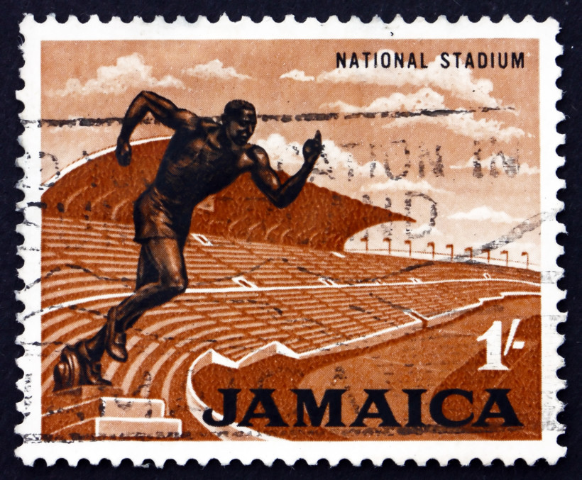 Jamaika Jamaikanische Briefmarke mit der Aufschrift 'National Stadium' und 'Jamaica' in Großbuchstaben. Abgebildet ist ein stilisierter Sprinter in dunkler Farbe, der aus den Zuschauerreihen eines großen Stadions auf eine Laufbahn sprintet. Der Hintergrund ist in