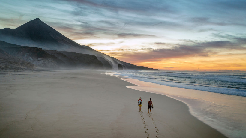 Zwei Personen gehen am Strand entlang, links Felsklippen, rechts Brandung bei Sonnenuntergang, Strandroute Jandia